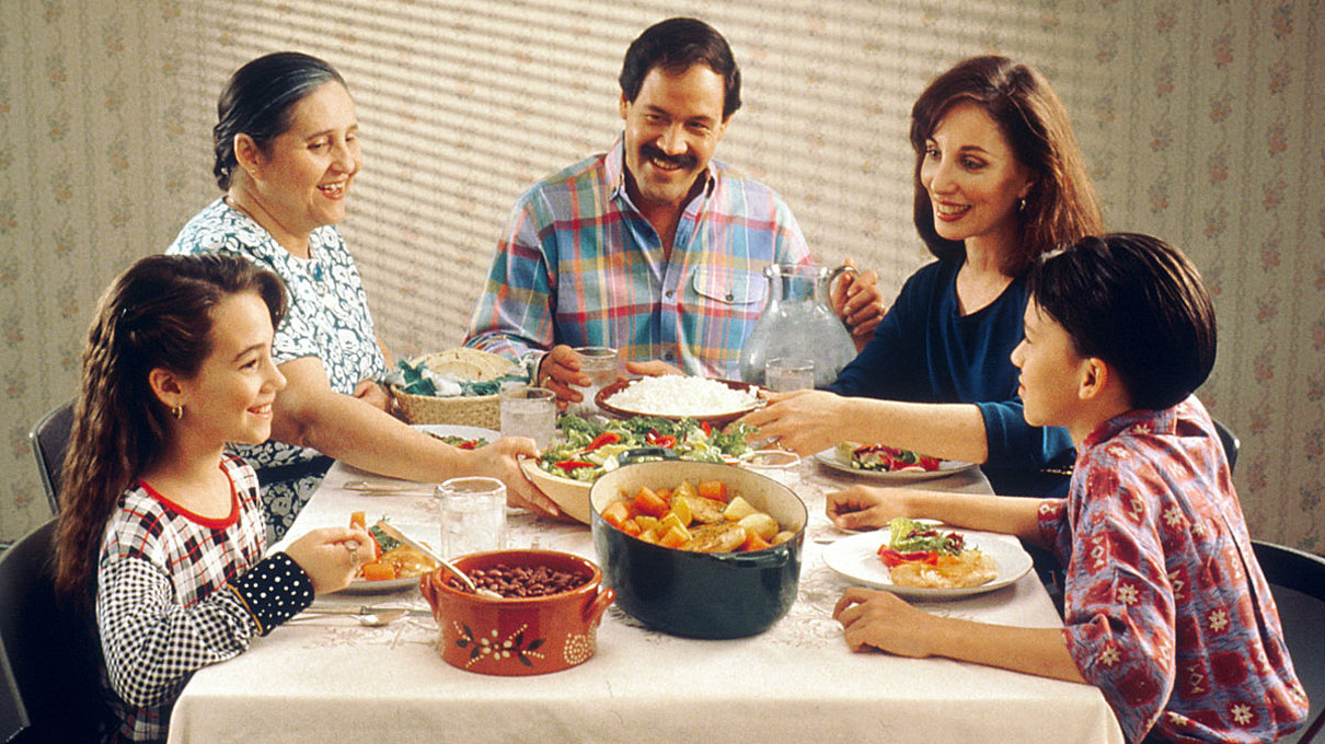 A Hispanic family (male adult, two female adults, female child, and male child) enjoy a meal at the dinner table.