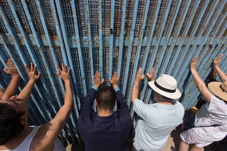 The Revs. Joel Hortiales (center, in blue blazer) and David Farley (to Hortiales' right) join parishioners of the Border Church in Tijuana, Mexico, as they lift their arms skyward beneath the fence that marks the border with the U.S. Photo by Mike DuBose, UMNS.