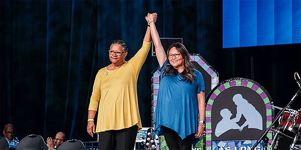 Sally Vonner, United Women in Faith top executive, and Bethany Amey Sutton, secretary of the group’s Executive Board, join hands on stage at the United Methodist General Conference in Charlotte, N.C., on April 29. Photo by Corbin Payne.