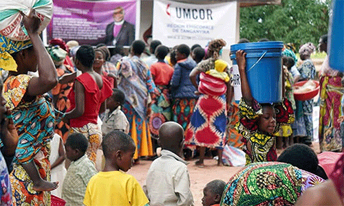 A young girl carries a bucket of supplies on her head amid a crowd of beneficiaries gathered in front of banners from UMCOR and the Tanganyika Episcopal Area. The distribution of relief kits aims to restore the dignity of families in Nyunzu, who are struggling to survive in the face of food insecurity and disease resulting from recent floods and conflicts in eastern DRC. Photo courtesy of the Disaster Management Office of the Tanganyika Episcopal Area.