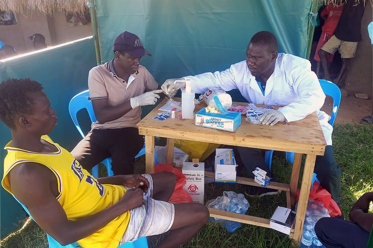 Arike Joel (à droite), Technicien de Laboratoire, réalise des dépistages du VIH lors d’un tournoi de football organisé par l’Église Méthodiste Unie au camp d’Imvepi, à Arua, en Ouganda. Ces dépistages s’inscrivent dans le cadre de la campagne « Kick Out HIV » menée par l’Église, qui met l’accent sur la prévention du VIH et la prise en charge personnelle dans les camps de réfugiés. Tandis que les joueurs se disputaient la victoire sur le terrain, des professionnels de santé proposaient, en marge du tournoi, des dépistages confidentiels du VIH et des consultations. Photo de Juma Denis Daniel, UM News.