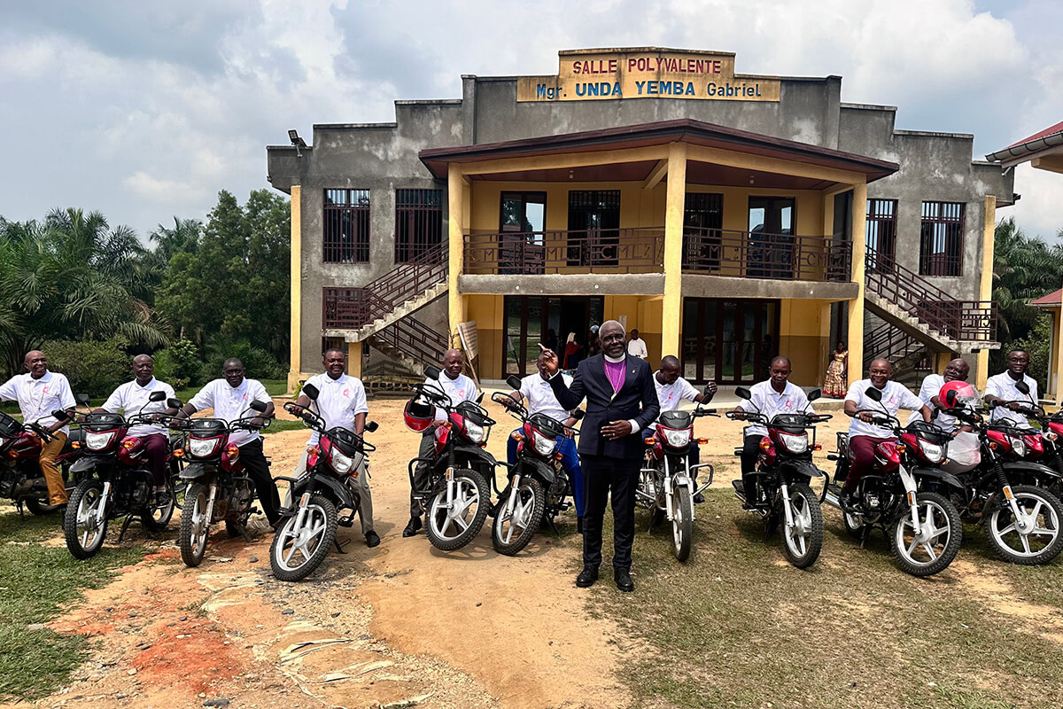 L'Evêque Antoine Tambwe Kalema (au centre) pose pour une photo aux côtés des 11 Surintendants de District de la Conférence du Congo Oriental qui ont reçu de nouvelles motos en cadeau de la part de l'Evêque lors de la Conférence Annuelle de 2026. Bon nombre de ces Responsables d'Église avaient passé des années à se déplacer à pied ou à vélo pour exercer leur ministère au sein de leurs communautés. Derrière eux se trouve la salle polyvalente Mgr Gabriel Unda Yemba, au Centre Mama Lynn de Kindu, au Congo. Photo de Chadrack Tambwe Londe, UM News.