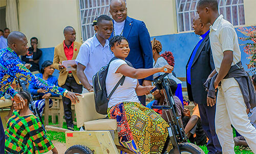 A young girl with limited mobility smiles as she maneuvers her tricycle equipped with hand cranks for the first time as Bishop Mande Muyombo (standing at center) and other church leaders look on. This project aims to promote social and educational inclusion by enabling young people and adults to travel with dignity to their places of work or study. Photo by Yamba Kisonga Barthélémy, UM News.
