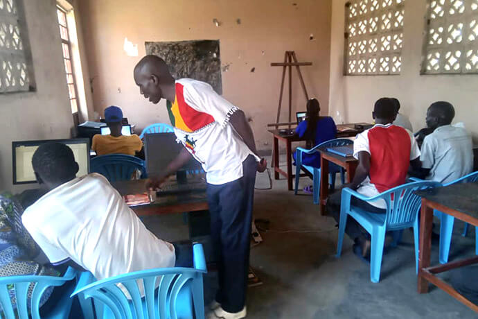 Hakim Erezenious Kwenyo (debout) aide un élève lors d'une séance du projet « Youth Digital Literacy » au centre de formation pour femmes de l'Église Méthodiste Unie d'Ariaze, situé dans le camp de Rhino, dans la zone de Siripi, en Ouganda. Ce projet bénéficie du soutien du ministère de l'Enfance du Soudan du Sud de l'Église Méthodiste Unie et de la Fondation Holston. Photo de Mudomo Joseph, UM News.